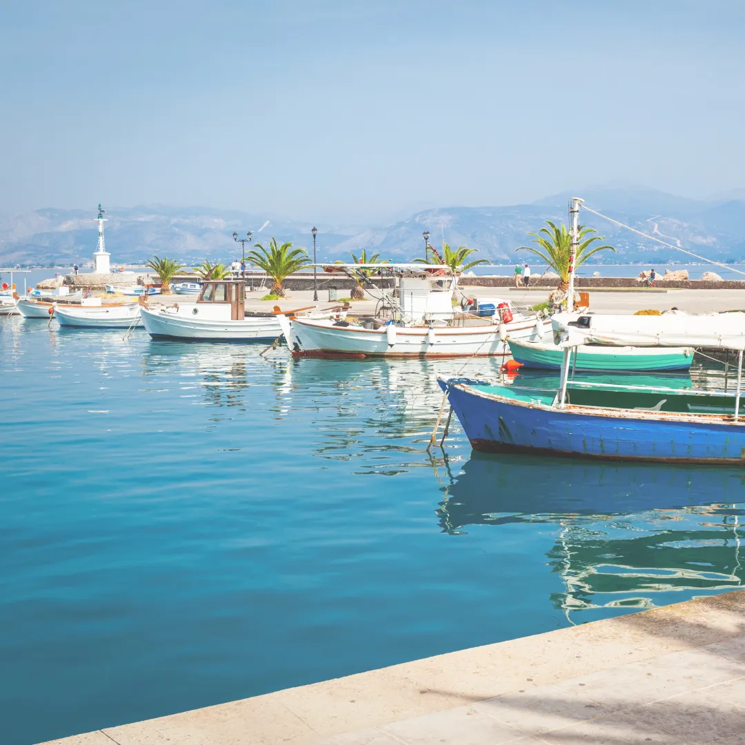 Ηλιόλουστη μέρα στο λιμάνι του Ναυπλίου με ψαρόβαρκες και τον φάρο στο βάθος, έτοιμο για βόλτα.