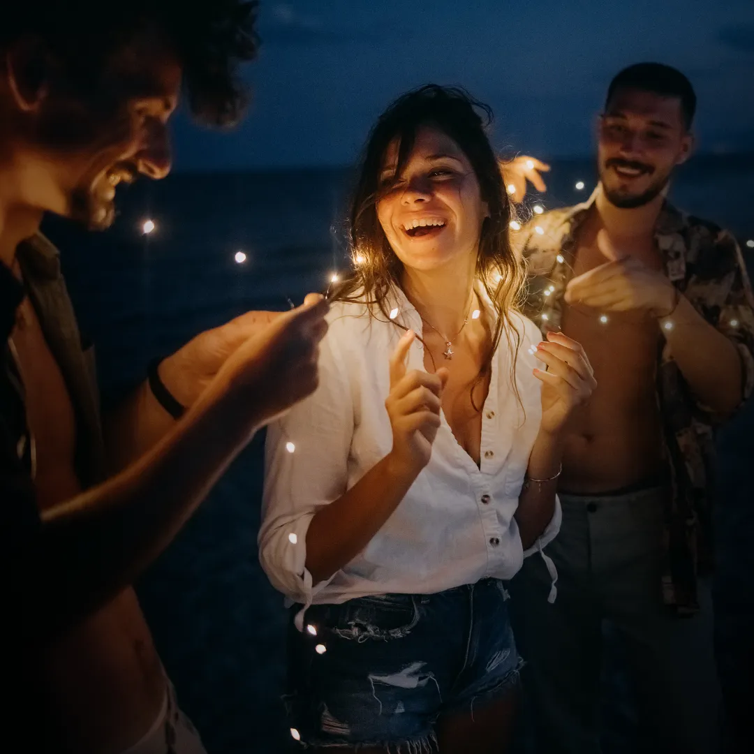 Παρέα φίλων που γελάει και διασκεδάζει τη νύχτα στην παραλία με λαμπάκια, έχοντας εξασφαλίσει VIP van για bachelor party στην Αθήνα.